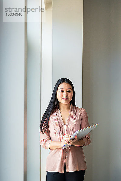 Porträt einer selbstbewussten Geschäftsfrau mit Akte  die im Büro an der Wand steht