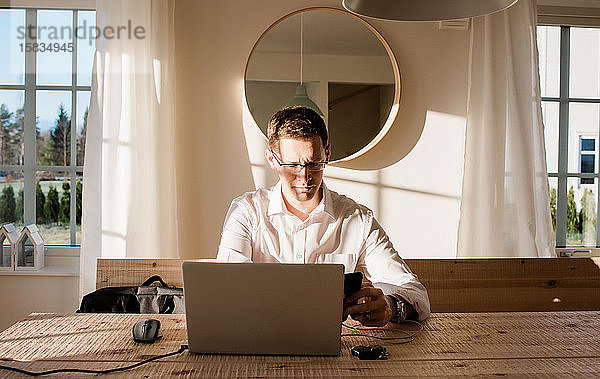 Mann arbeitet von zu Hause aus und schaut konzentriert auf sein Telefon und seinen Laptop