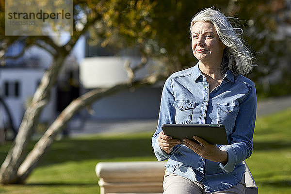 Nachdenkliche Frau  die einen Tablet-Computer hält  während sie an einer Stützmauer im Park an einem sonnigen Tag steht