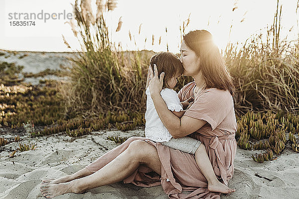 Seitenansicht einer Mutter  die ein junges Kleinkind bei Sonnenuntergang am Strand küsst