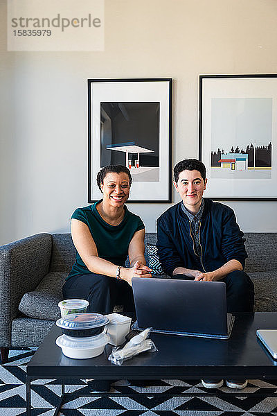 Porträt von selbstbewusst lächelnden Geschäftskollegen mit Laptop am Tisch sitzend im Büro