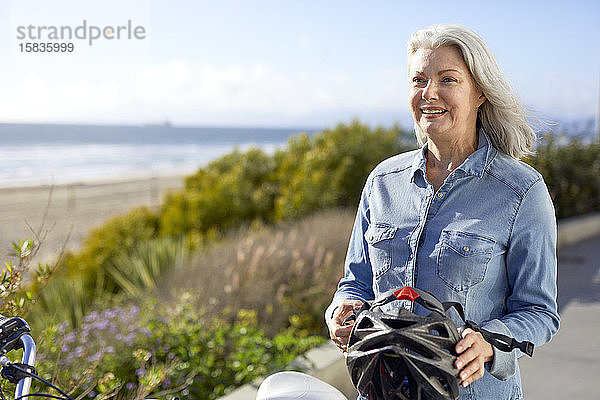 Lächelnde ältere Frau mit Fahrradhelm  während sie an einem sonnigen Tag auf der Straße gegen den Himmel steht
