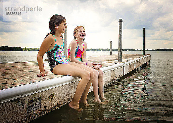 Zwei junge Mädchen in Badeanzügen sitzen lachend auf einem Dock am See