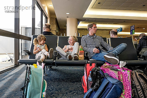 zwei Mädchen und ein Vater sitzen lachend im Flughafenterminal und warten