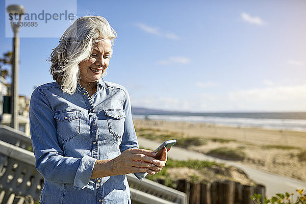 Lächelnde ältere Frau benutzt Smartphone  während sie am Strand von Manhattan an einem sonnigen Tag gegen den Himmel steht