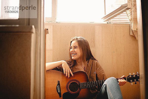 Fröhliche junge Frau hält Gitarre  während sie auf dem Balkonboden sitzt