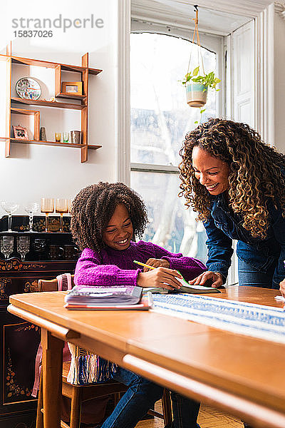 Lächelnde Mutter hilft Tochter bei den Hausaufgaben bei Tisch im Wohnzimmer
