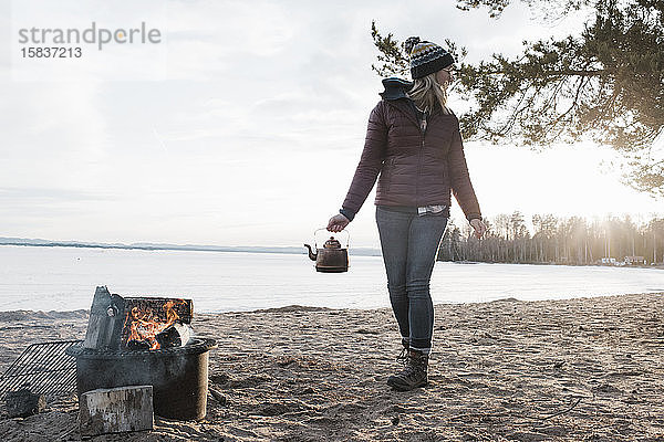 Frau  die in Schweden mit einem Teekessel zum Lagerfeuer an einem Strand geht