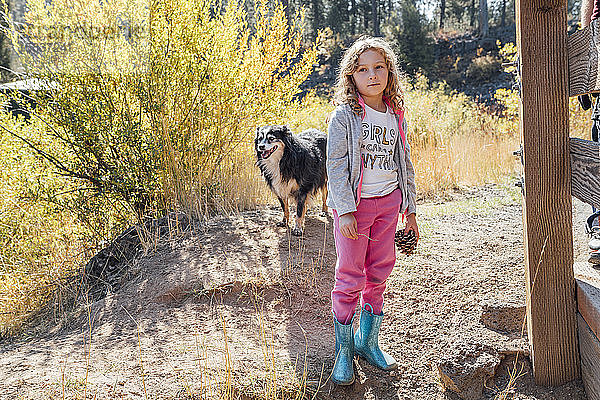 Mädchen hält Tannenzapfen und steht im Herbst mit Hund im Feld