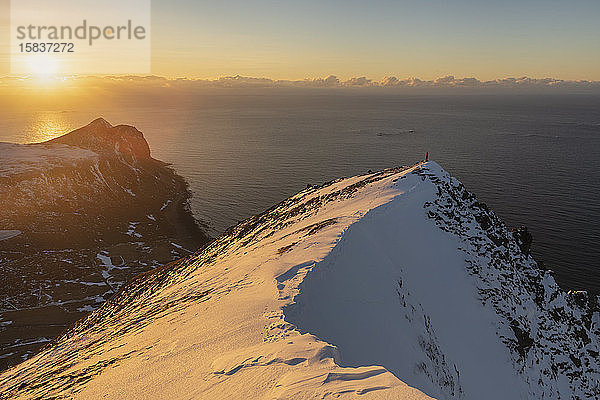 Wanderin steht allein auf schneebedecktem Berggipfel mit der Sonne im Hintergrund  FlakstadÃ¸y  Lofoten  Norwegen