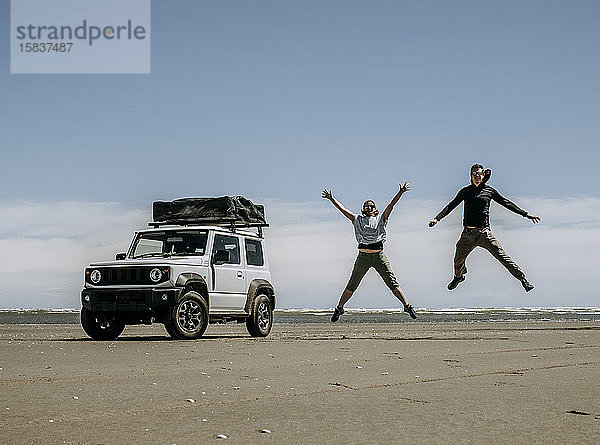 Paar springt vor Freude neben seinem am Strand in Neuseeland geparkten Geländewagen