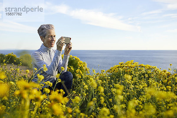 Ältere Frau fotografiert mit einem Smartphone  während sie sich bei Pflanzen gegen den Himmel hockt