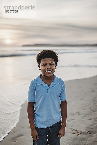 Porträt eines jungen Jungen im Schulalter  der bei Sonnenuntergang am Strand lächelt