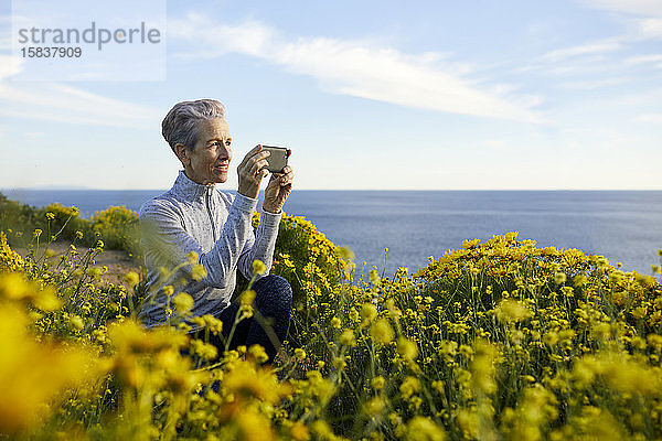 Ältere Frau fotografiert mit einem Smartphone  während sie am Meer vor Pflanzen gegen den Himmel kauert