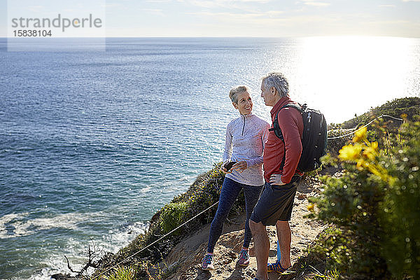 In voller Länge lächelndes älteres Ehepaar redet  während es an einem sonnigen Tag auf einer Klippe am Meer steht