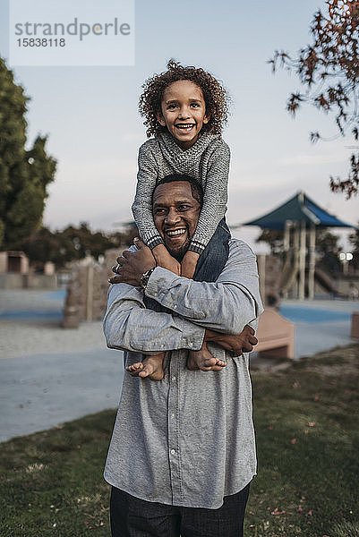 Fröhlicher Sohn sitzt auf den Schultern des glücklichen Vaters auf einem Spielplatz im Park