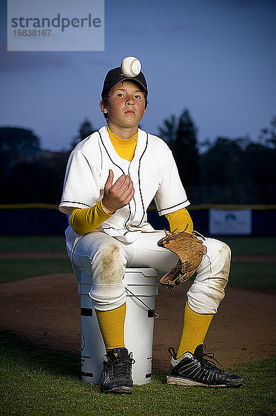 Porträt eines Jungen in Baseball-Uniform  der auf einem Eimer sitzen und Baseball werfen soll