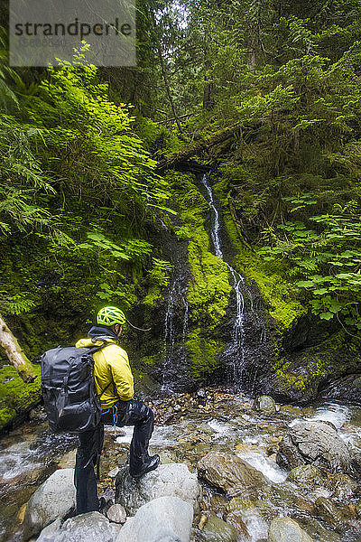 nasser Wanderer mit Rucksack schaut auf kleinen Wasserfall.