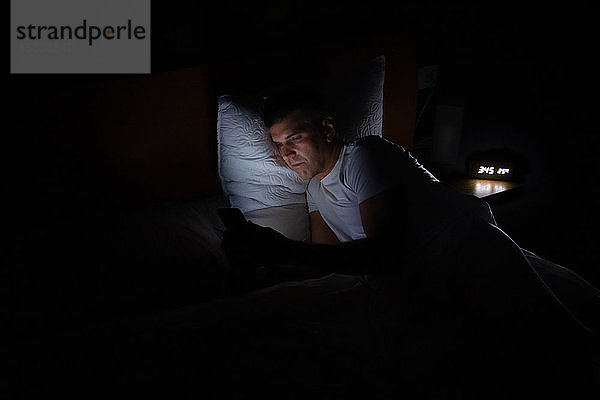 Ein Mann liegt in völliger Dunkelheit im Bett und schaut auf sein Smartphone