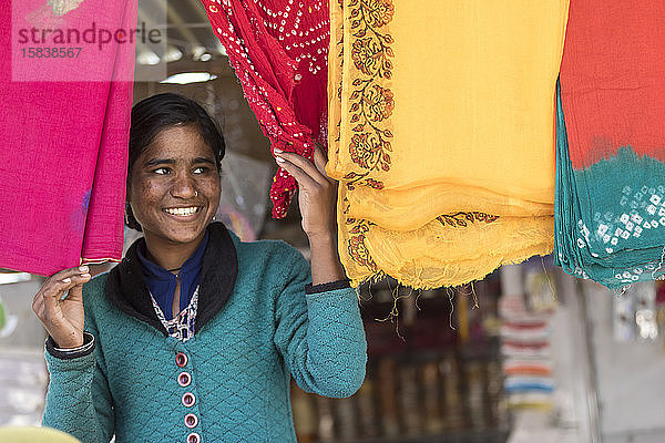 Eine junge Frau blickt zwischen bunten indischen Kleidern hervor und lächelt.
