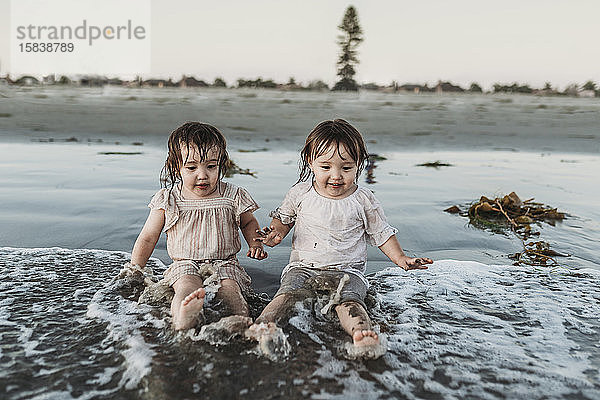 Frontansicht der kleinkindigen Schwestern  die am Strand sitzen und im Wasser planschen