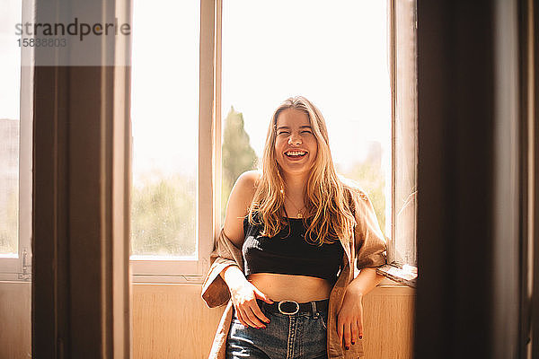 Fröhliche junge Frau lacht  während sie im Sommer auf dem Balkon steht