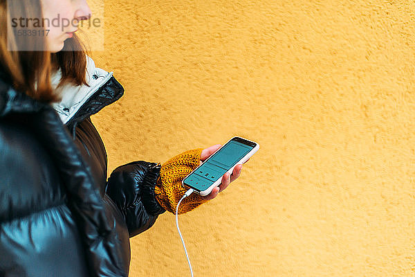 Junge Frau benutzt Smartphone  während sie an gelber Wand steht