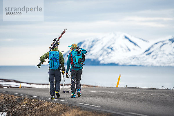 Ein Ehepaar geht in Island mit Skiausrüstung eine Straße entlang