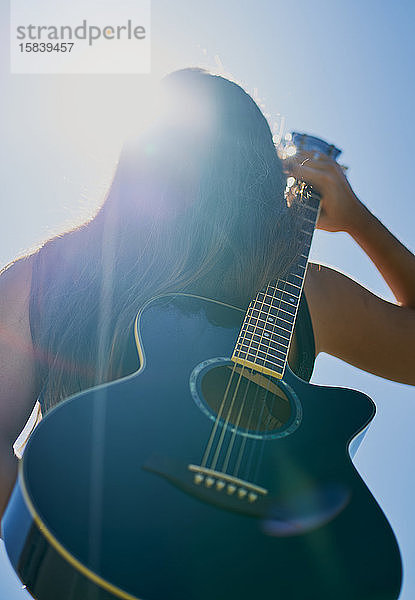 Eine weibliche Teenager-Gitarristin hält sich eine Akustikgitarre auf dem Rücken vor einen blauen Himmel