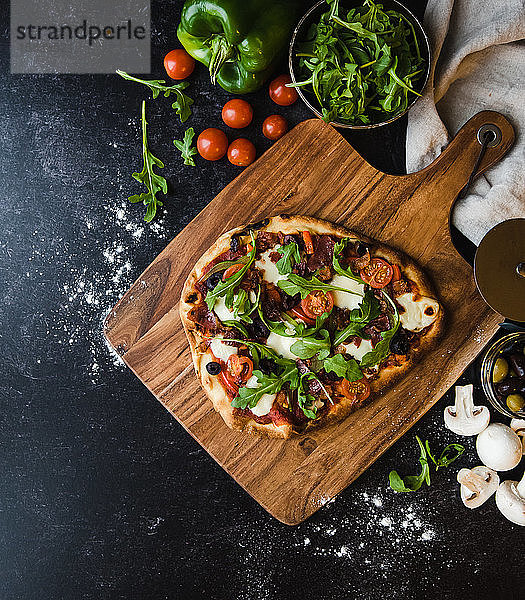 Draufsicht auf eine handgemachte Pizza auf Holzbrett mit Zutaten drum herum.