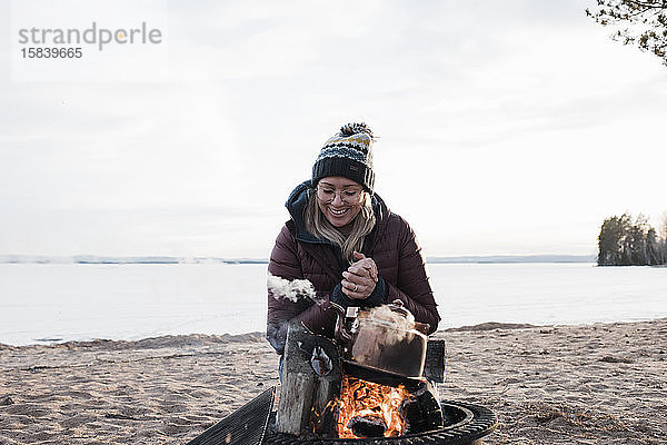 Frau  die sich im Winter am Strand am Lagerfeuer wärmt und lächelt