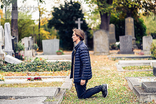 Trauernder junger Mann kniend vor einem Grab auf einem Friedhof