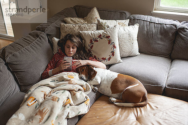 Tweener-Mädchen schaut auf ihr Telefon  während sie mit dem Jagdhund auf der Couch kuschelt