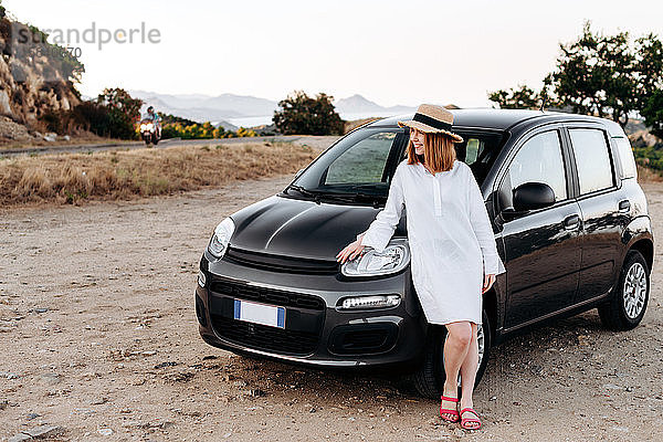 Ein süßes  charmantes Mädchen mit Strohhut posiert um ein schwarzes Auto am Straßenrand.