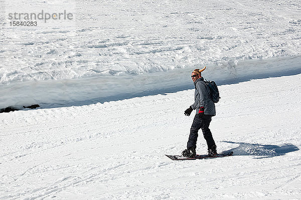Snowboarder  der im Skigebiet mit einer lustigen Mütze den Hang hinunter rennt. Wintersport und Erholung  Freizeitaktivitäten im Freien