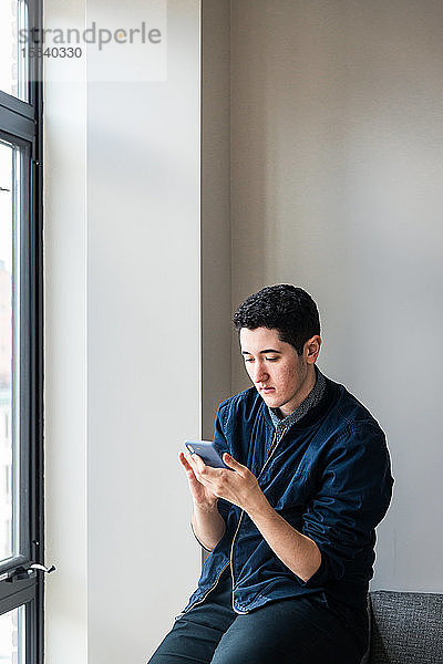 Junger Geschäftsmann benutzt Smartphone  während er im Büro auf dem Sofa an der Wand sitzt