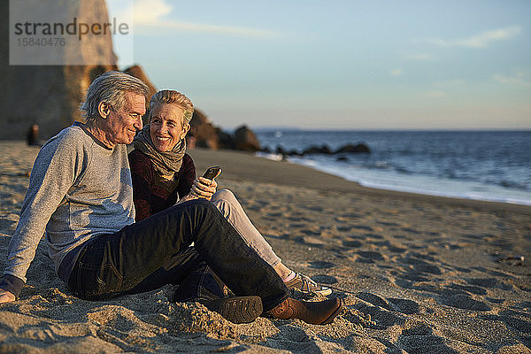 Älteres Ehepaar telefoniert  während es bei Sonnenuntergang am Strand im Sand sitzt