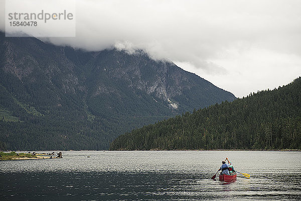 Mann und Kind paddeln mit einem roten Kanu auf einem Bergsee an einem bewölkten Tag