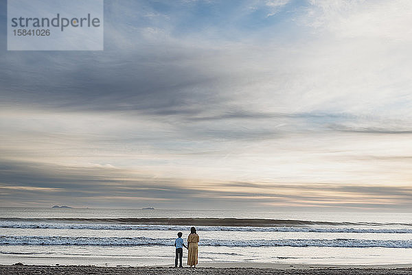 Junge Mutter mit Kleid und Sohn stehen bei bewölktem Sonnenuntergang am Strand