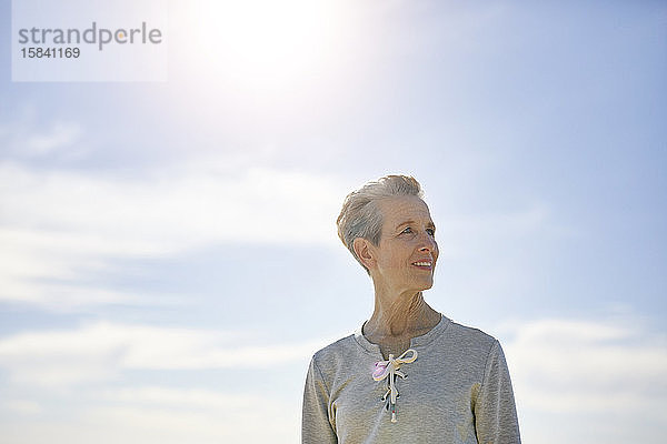 Lächelnde ältere Frau schaut weg  während sie an einem sonnigen Tag am Himmel steht