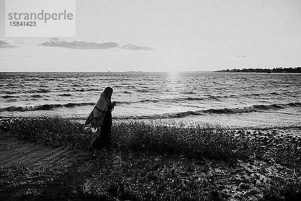 Frau  die bei Sonnenuntergang in einer Decke am Strand spazieren geht