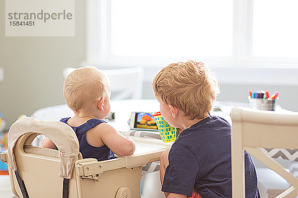 Rückansicht eines Mädchens und eines kleinen Bruders beim Telefonieren am Tisch im Haus