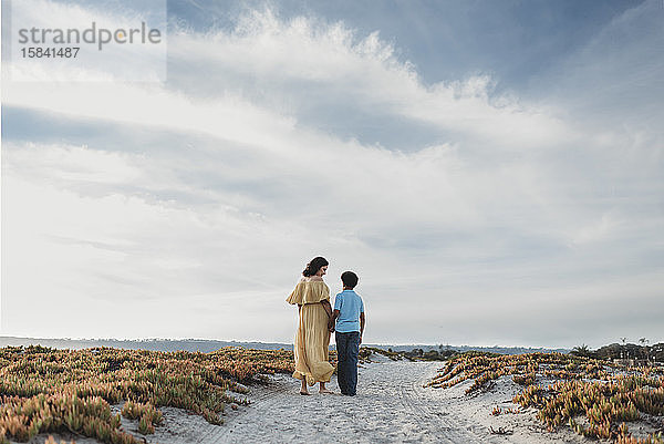 Hinteransicht von Mutter und Sohn beim Spaziergang am Strand vor wolkenblauem Himmel