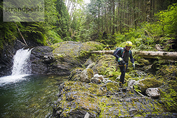 Wanderer wandert über felsiges Gelände neben einem Wasserfall in dichtem Wald.