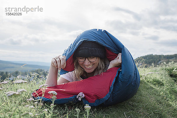 Porträt einer Frau  die beim Zelten auf einem Hügel in einem Schlafsack lacht