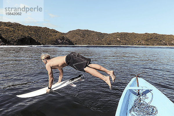 Mann ohne Hemd mit Surfbrett  der bei Sonnenschein gegen den Himmel ins Meer springt