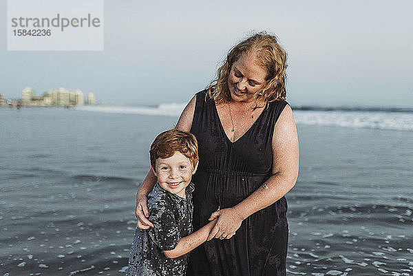 Porträt eines lächelnden Sohnes mit Mutter in der Abenddämmerung im Meer am Strand