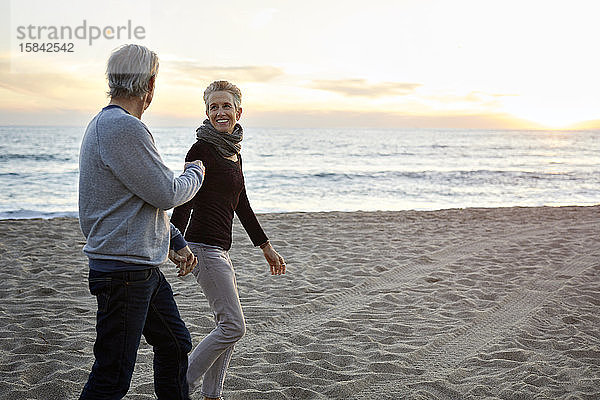 Lächelndes älteres Paar hält sich beim Strandspaziergang bei Sonnenuntergang an den Händen