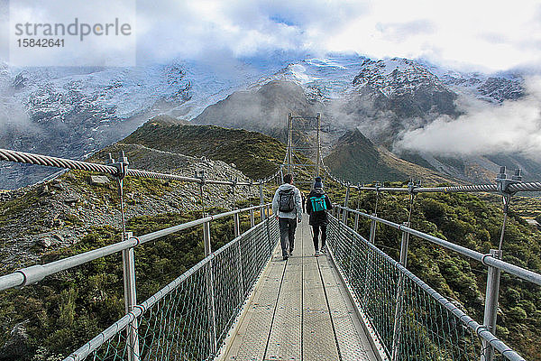 Mann und Frau gehen auf einer Hängebrücke  die von Bergen umgeben ist.