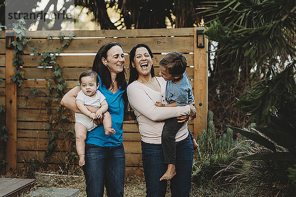 Lachende Familie mit zwei Müttern und zwei Kindern im Hinterhof beim Zaun